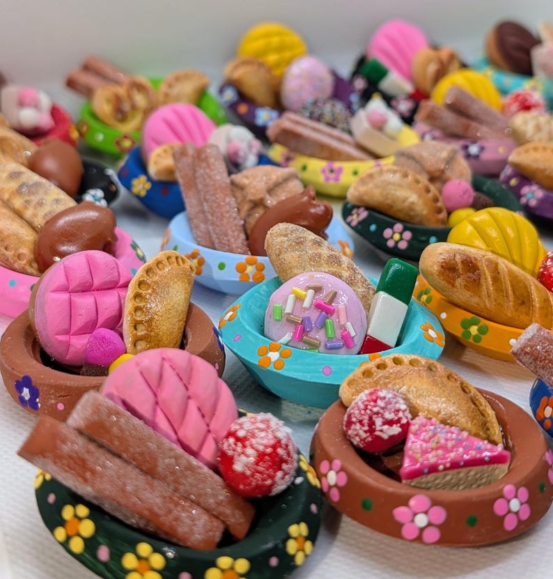 Może przedstawiać: Kolekcja miniaturowych glinianych miseczek, ręcznie malowanych w kwiatowe wzory, zawierających r&oacute;żnorodne rzeźbione produkty spożywcze, w tym małe pieczywo, wypieki, ciasteczka oraz kolorowe słodycze, takie jak r&oacute;żowe conchas i posypane cukrem smakołyki.