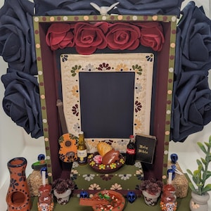 May include: A decorative Day of the Dead altar with a dark blue frame and dark blue fabric roses. The altar features a central frame, miniature food items, a guitar, and a miniature Holy Bible. The altar is decorated with colorful details and floral patterns.