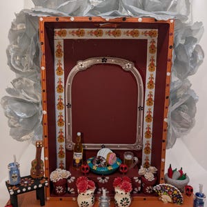 May include: A Day of the Dead-themed shadow box with a red interior and silver frame. The box is decorated with miniature guitars, skulls, flowers, and food items. Silver fabric flowers adorn the top. A Corona beer bottle and a beer mug are also visible.