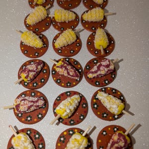 May include: A collection of 18 miniature corn on the cob, some with yellow kernels and white cheese, others with red and white stripes, all on small brown plates with white and yellow flower designs.