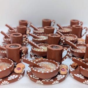 May include: A collection of miniature brown ceramic bowls, mugs, spoons, and plates, each with white floral detailing. The bowls contain a light-colored liquid, and some plates have small cookies and cinnamon sticks. The items are arranged on a white surface.