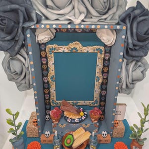 May include: A Day of the Dead altar with a teal frame, adorned with gray roses and decorative elements. The altar features miniature food items, skulls, and floral accents, creating a festive and colorful display.