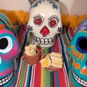 May include: A Day of the Dead themed display featuring a central white skull with red accents and a floral design. Flanking the skull are two colorful skull figurines. The display includes miniature food items and is set on a striped textile.