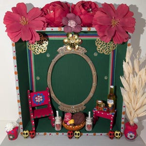 May include: A Day of the Dead altar with a green background, a gold framed mirror, a red chair, a small table, a bottle of Corona beer, a bowl of pan dulce, and sugar skulls. The altar is decorated with red flowers, gold paper flowers, and a dried wheat stalk.