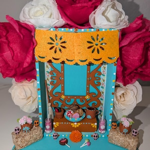 May include: A Day of the Dead altar with a turquoise blue background, a brown frame, and a yellow paper banner. The altar is decorated with flowers, skulls, candles, and other traditional Mexican decorations.