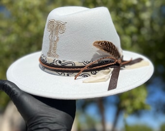 Pode incluir: Chapéu fedora branco com uma bandana paisley marrom, uma pena marrom e uma letra "I" bordada em ouro na frente.
