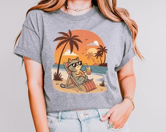 May include: A gray t-shirt with a cartoon cat wearing sunglasses and holding a drink, relaxing on a beach chair under palm trees with a sunset in the background.