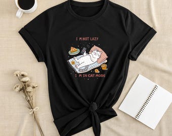 May include: Black t-shirt with a graphic of a white cat lounging on a rug, holding a phone, and surrounded by food. The text "I'M NOT LAZY I'M IN CAT MODE" is above and below the cat. A cup of coffee and a notebook are also visible.
