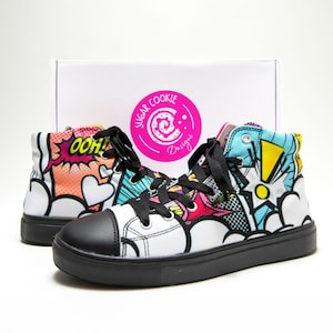 Puede incluir: Zapatillas altas blancas con suelas y cordones negros. Las zapatillas presentan un diseño colorido de estilo cómic con la palabra "OOH!" y otros elementos gráficos. Una caja blanca con un logotipo rosa está en el fondo.
