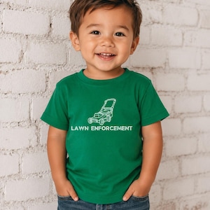 Op de afbeelding: Een groen T-shirt met korte mouwen en een witte lijntekening van een grasmaaier in het midden op de borst. Onder de afbeelding van de grasmaaier staat de tekst LAWN ENFORCEMENT gedrukt in een vet, wit, schreefloos lettertype.