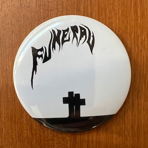May include: A white button with a black and white image of a cross and the word "FUNERAL" in a gothic font.