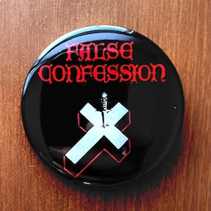 May include: A round, black button with the words "FALSE CONFESSION" in red, gothic-style lettering. A white cross with a red outline and a sword through it is centered below the text. The button has a glossy finish.