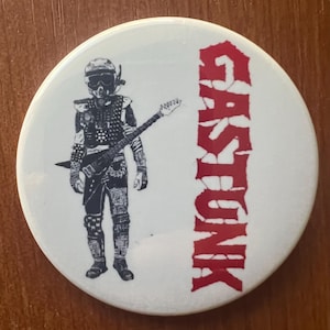 May include: A white button with a black and white illustration of a person wearing a helmet and holding a guitar. The word "GASTUNK" is written in red letters on the right side of the button.