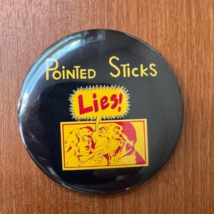May include: Black button with yellow text that reads "Pointed Sticks Lies!" and a cartoon image of two people. The image shows a woman with blonde hair and a man with a beard. The woman is pointing at the man with her right hand.