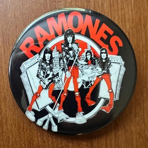 May include: Round black button featuring the band name "RAMONES" in red block letters. The button has a graphic of the band members in black and white, with red pants, playing instruments in front of a city skyline.