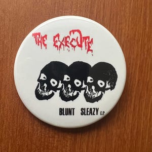May include: A white button with the text "THE EXECUTE" in red, above three black skull illustrations. Below the skulls is the text "BLUNT SLEAZY E.P." in black. The button is round and sits on a wooden surface.
