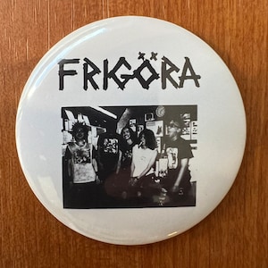 May include: A white button with a black and white photo of a band called "FRIGÖRA" on it. The band members are standing in a room with a few posters on the wall.