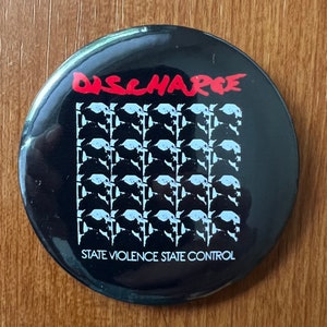 May include: A black and white button badge with the band name "DISCHARGE" in red letters and the text "STATE VIOLENCE STATE CONTROL" in white letters.