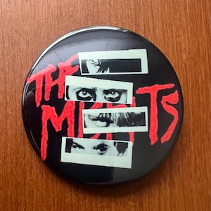 May include: A round, black button with the words "THE MIST" in red, stylized lettering. The button features four rectangular black and white images of eyes and mouths.