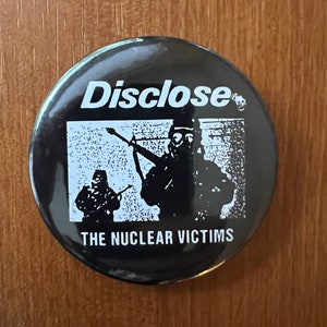 May include: Black and white button badge with the text "Disclose" and "The Nuclear Victims" in white lettering. The image shows two figures in gas masks holding rifles.
