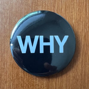 May include: A round, black button with the word "WHY" in large, light blue capital letters. The button has a glossy finish and is set against a wooden surface.