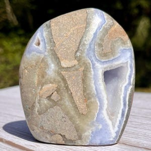 May include: A polished, freeform stone with a geode cavity. The stone features a mix of tan, gray, and pale blue colors, with a visible crystalline structure. The geode opening reveals a contrasting white interior, creating a natural, decorative piece.