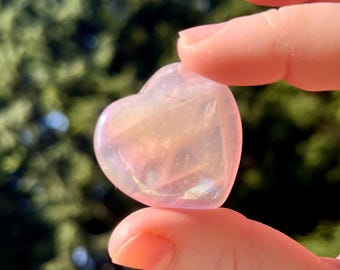 May include: A heart-shaped, iridescent crystal in shades of pink and peach. The crystal is held between two fingers, with a blurred green background of foliage. The crystal's surface reflects light, creating a shimmering effect.