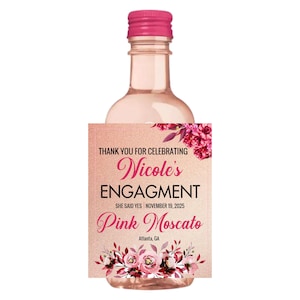 May include: A small pink Moscato wine bottle with a pink cap. The label reads "Nicole's ENGAGMENT" with the text "SHE SAID YES NOVEMBER 19, 2025" and "Pink Moscato". Floral accents adorn the label.