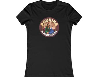 May include: Black fitted t-shirt featuring a circular graphic with the words "Pouring & Parenting." The graphic depicts two women, one in a green dress and the other in a blue dress, with a gold-colored background.