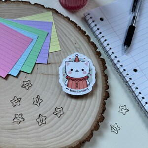 May include: A white cat wearing a red and white clown costume with a party hat. The sticker says "My brain is a circus".