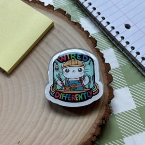 May include: A white cat wearing a yellow hard hat and blue overalls is holding a pair of pliers and a wire. The cat is surrounded by colorful wires and the text "Wired Differently".