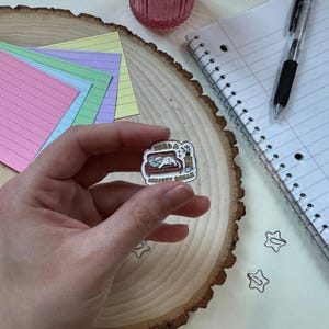 Puede incluir: Un pin de esmalte blanco con un gato de dibujos animados en un sofá. El gato lleva una camisa blanca y tiene una planta a su lado. El texto en el pin dice "Need a Sensory Break".