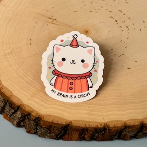 May include: A white cat wearing a red and orange clown costume with a red and white hat. The cat has a cute expression on its face. The sticker says "My brain is a circus".