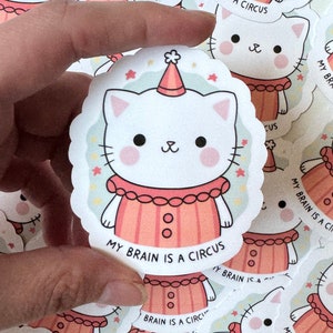 May include: A white cat wearing a red and white clown costume with a party hat. The cat has pink cheeks and black eyes. The text "My brain is a circus" is written below the cat.