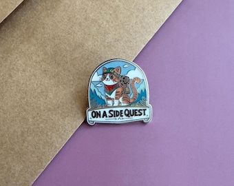 Peut inclure: Épingle en émail représentant un chat aventurier de dessin animé avec un sac à dos, un bandana et des lunettes, sur fond de montagnes. L'épingle porte le texte "ON A SIDE QUEST" en bas, avec un code couleur bleu clair et blanc.