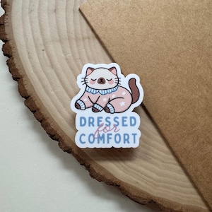 May include: A sticker featuring a cartoon cat in a pink outfit with white stars. The cat is wearing a blue collar and the text "Dressed for Comfort" is below the cat in blue and pink.
