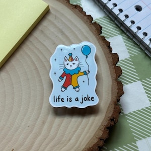 May include: A sticker featuring a cartoon cat dressed as a clown, holding a blue balloon. The cat wears a party hat and a red, yellow, and blue outfit. The text "life is a joke" is written below the cat on a light blue background.