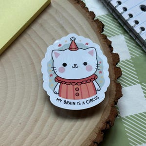 May include: White cat wearing a red and white striped clown costume with a party hat. The cat has pink cheeks and a cute expression. The sticker says "My brain is a circus".