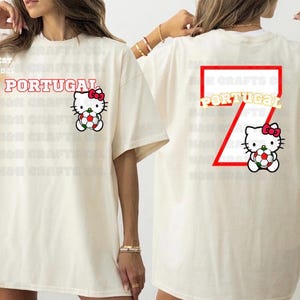 K&ouml;nnte beinhalten: Cremefarbenes T-Shirt mit dem Wort "PORTUGAL" in Rot, einem Hello Kitty-Motiv mit einem Fu&szlig;ball und der Zahl "7" auf dem R&uuml;cken. Das Shirt hat eine lockere Passform und kurze &Auml;rmel. Der Text "EST 192" ist ebenfalls sichtbar.