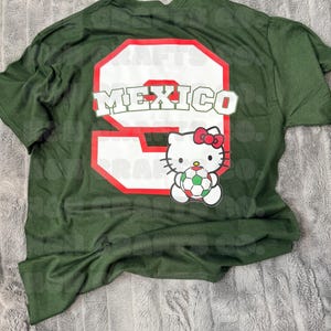K&ouml;nnte beinhalten: Dunkelgr&uuml;nes T-Shirt mit einer gro&szlig;en wei&szlig;en Zahl 9 mit roter Umrandung und dem Wort "MEXICO" in Wei&szlig;. Darunter eine Hello Kitty-Grafik, die einen Fu&szlig;ball h&auml;lt. Das Shirt liegt auf einer grau strukturierten Oberfl&auml;che.