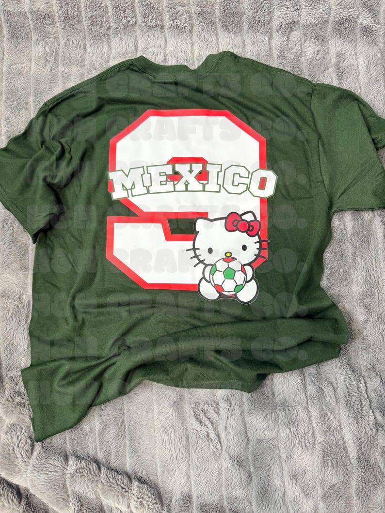 K&ouml;nnte beinhalten: Dunkelgr&uuml;nes T-Shirt mit einer gro&szlig;en wei&szlig;en Zahl 9 mit roter Umrandung und dem Wort "MEXICO" in Wei&szlig;. Darunter eine Hello Kitty-Grafik, die einen Fu&szlig;ball h&auml;lt. Das Shirt liegt auf einer grau strukturierten Oberfl&auml;che.