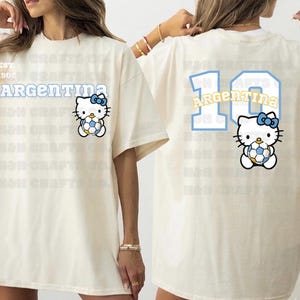 K&ouml;nnte beinhalten: Cremefarbenes T-Shirt mit einem Hello Kitty-Design, das einen Fu&szlig;ball h&auml;lt, mit dem Wort "ARGENTINA" dar&uuml;ber. Auf der R&uuml;ckseite des Shirts sind die Nummer "10" und das Wort "ARGENTINA" in &auml;hnlichem Stil sowie das Hello Kitty-Design abgebildet.