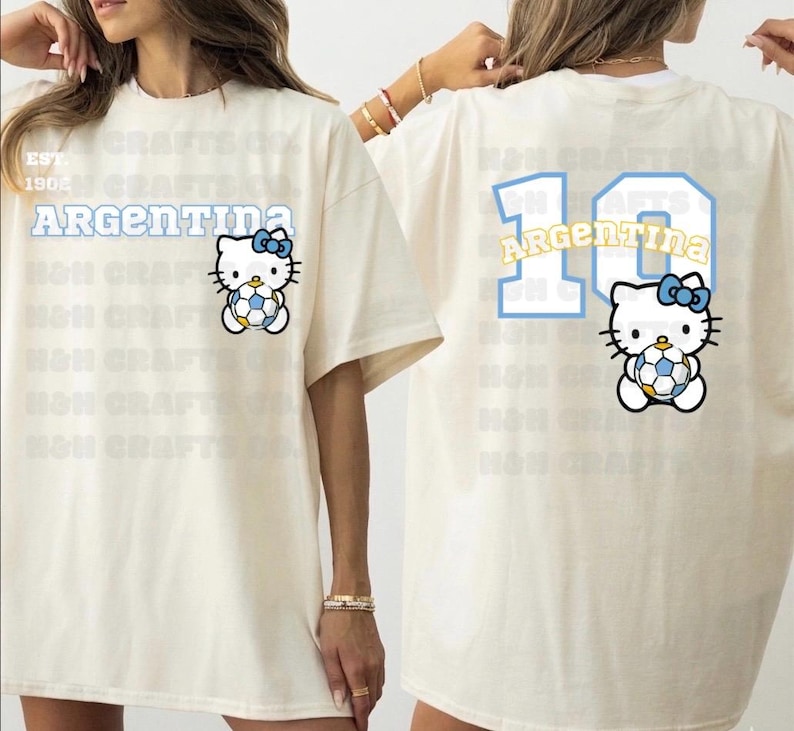 K&ouml;nnte beinhalten: Cremefarbenes T-Shirt mit einem Hello Kitty-Design, das einen Fu&szlig;ball h&auml;lt, mit dem Wort "ARGENTINA" dar&uuml;ber. Auf der R&uuml;ckseite des Shirts sind die Nummer "10" und das Wort "ARGENTINA" in &auml;hnlichem Stil sowie das Hello Kitty-Design abgebildet.