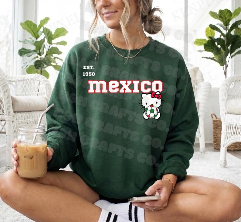 K&ouml;nnte beinhalten: Ein dunkelgr&uuml;nes Sweatshirt mit dem Wort "mexico" in roter und wei&szlig;er Schrift. Das Sweatshirt zeigt auch eine Hello Kitty-Grafik, die einen Fu&szlig;ball h&auml;lt. Der Text "EST. 1950" steht &uuml;ber dem Wort "mexico".