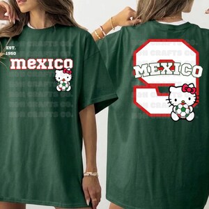 K&ouml;nnte beinhalten: Dunkelgr&uuml;nes T-Shirt mit dem Wort "MEXICO" in Rot und Wei&szlig; und einer Hello Kitty-Grafik, die einen Fu&szlig;ball h&auml;lt. Auf der R&uuml;ckseite befindet sich eine gro&szlig;e 9 mit "MEXICO" und eine weitere Hello Kitty-Grafik.