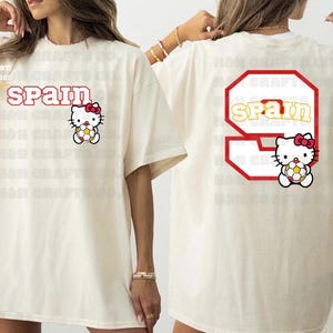 K&ouml;nnte beinhalten: Cremefarbenes T-Shirt mit dem Wort "SPAIN" in roter Schrift und einem Hello Kitty-Charakter, der einen Fu&szlig;ball h&auml;lt. Auf der R&uuml;ckseite des Shirts befindet sich eine gro&szlig;e 9 mit "SPAIN" darin und eine weitere Hello Kitty.