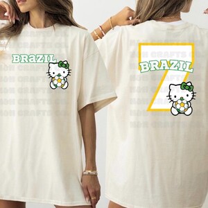 K&ouml;nnte beinhalten: Cremefarbenes T-Shirt mit dem Wort "BRAZIL" in Gr&uuml;n und einer Zeichentrickfigur, die einen Fu&szlig;ball h&auml;lt. Auf der R&uuml;ckseite des Shirts befindet sich ein gro&szlig;es gelb-gr&uuml;nes Design mit dem Wort "BRAZIL" und derselben Figur.