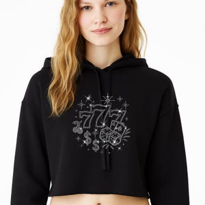 May include: Black cropped hoodie featuring a rhinestone design of casino symbols, including lucky sevens, cherries, dice, and dollar signs. The design is embellished with sparkling star accents, creating a glamorous look.