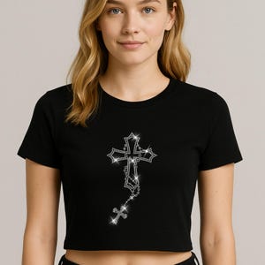 May include: Black crop top featuring a sparkling rhinestone cross design. The cross is detailed with a rosary-style chain and a smaller cross at the bottom. The shirt has short sleeves and a crew neck.