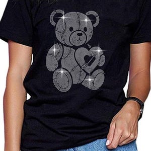 Puede incluir: Camiseta negra con un diseño de osito de peluche de strass que sostiene un corazón. El osito está hecho de strass brillantes.
